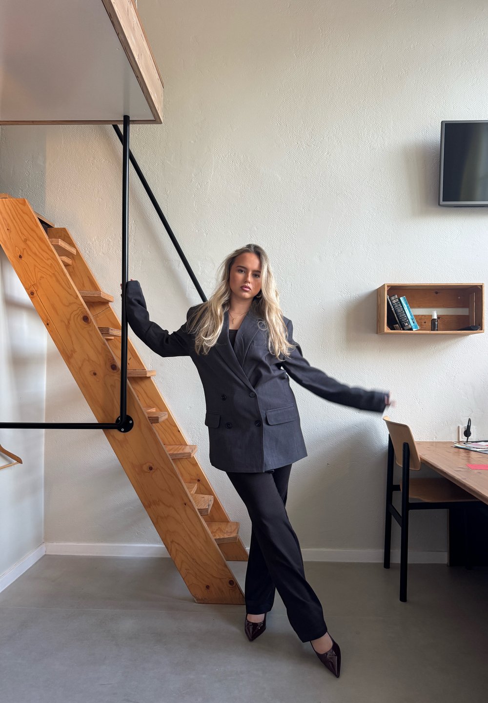 Blazer pe ton cenușiu închis, ajustat, cu nasturi dubli, asortat cu pantaloni strâmți de culoare închisă. Scări de lemn pentru cumpărături și decor minimalișt în fundal.