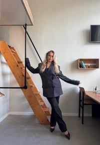 Blazer pe ton cenușiu închis, ajustat, cu nasturi dubli, asortat cu pantaloni strâmți de culoare închisă. Scări de lemn pentru cumpărături și decor minimalișt în fundal.