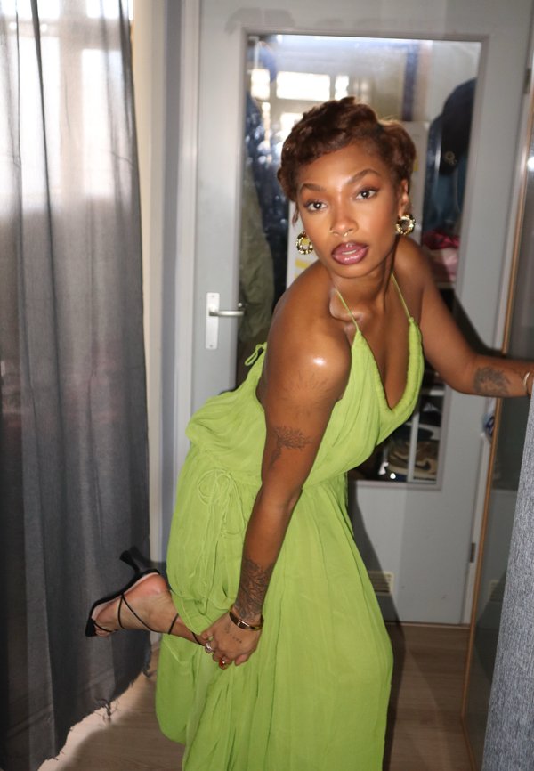 Γυναίκα με πράσινο φόρεμα halter που προσαρμόζει το μαύρο πέδιλο με λουράκια σε ένα δωμάτιο με γκρι κουρτίνες και μια λευκή πόρτα με καθρέφτη.