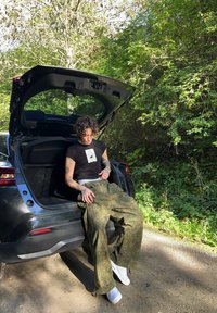 Juodas grafiškas marškinėliai su apvaliu kaklu, laisvi žali raštuoti kelnės ir baltos sportbačiai. Asmuo sėdi automobilio bagažinėje.