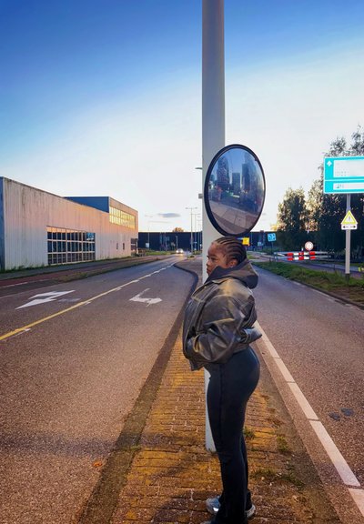 Espejo convexo redondo montado en un poste cerca de una carretera, reflejando edificios y árboles. Cerca, una persona con una chaqueta de cuero gris y pantalones negros está de pie.