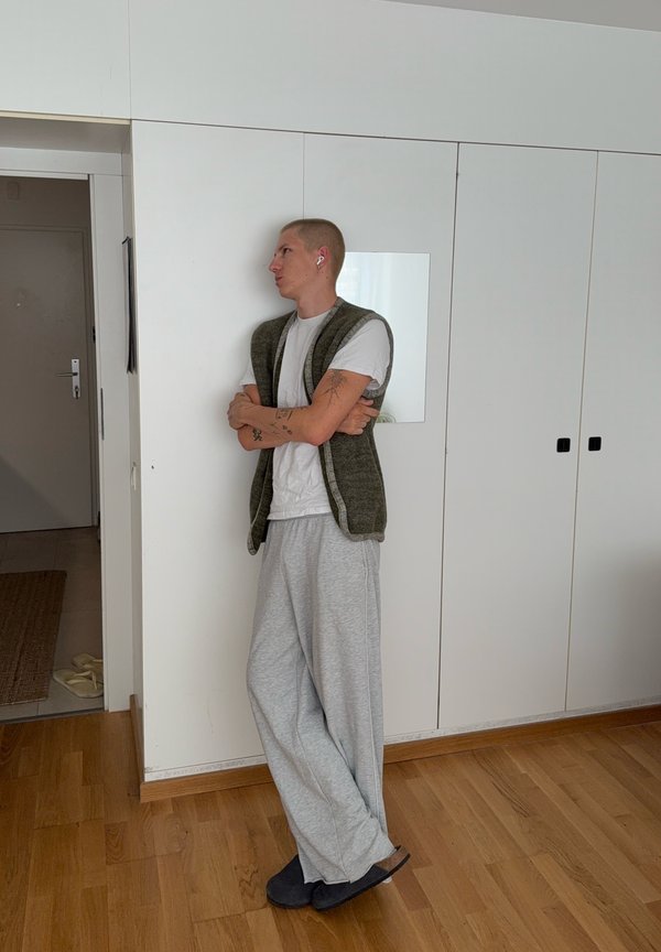 Persona con capelli corti che indossa auricolari, maglietta bianca, pantaloni della tuta grigi e un gilet verde, in piedi con le braccia incrociate davanti a armadi bianchi all'interno.