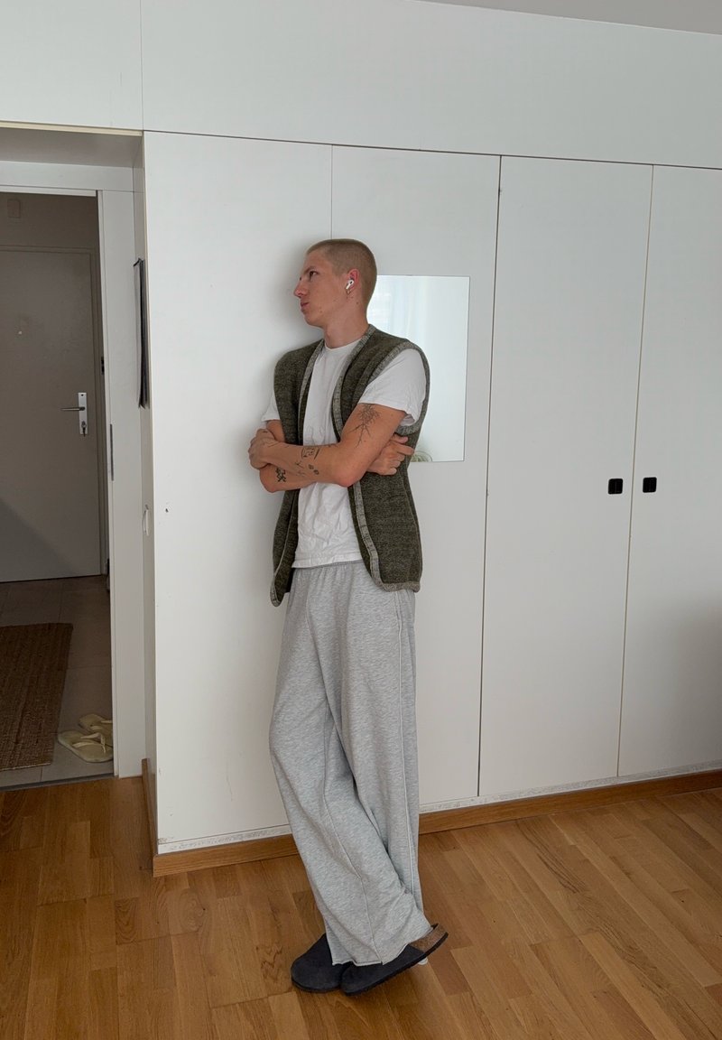 Personne aux cheveux courts portant des écouteurs, un t-shirt blanc, un pantalon de survêtement gris et un gilet vert, debout les bras croisés devant des armoires blanches à l'intérieur.