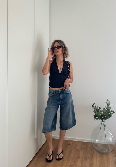 Mujer con top halter negro, culottes vaqueros, sandalias negras y gafas de sol, de pie junto a una pared blanca y un gran jarrón transparente con hojas verdes.
