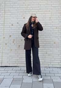 Femme en pantalon noir à jambes larges, manteau marron foncé, casquette beige, écouteurs autour du cou, debout contre un mur de briques clair, touchant sa casquette.