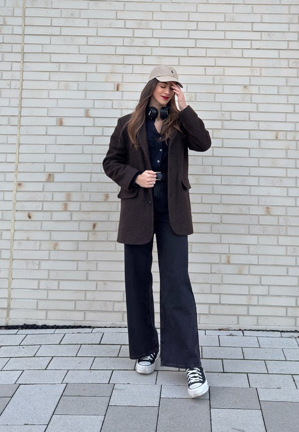 Femme en pantalon noir à jambes larges, manteau marron foncé, casquette beige, écouteurs autour du cou, debout contre un mur de briques clair, touchant sa casquette.
