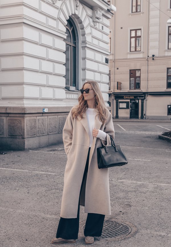 Woman wearing beige coat and sunglasses, holding black handbag, standing on city street near ornately designed stone building with arched window.