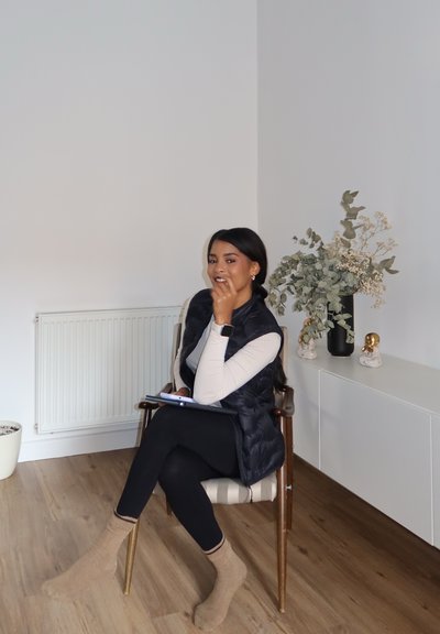 Chaleco negro de plumas sobre una blusa beige clara, leggings negros y calcetines color tostado. Sentada en una silla de madera con un cojín a rayas, sosteniendo un cuaderno.