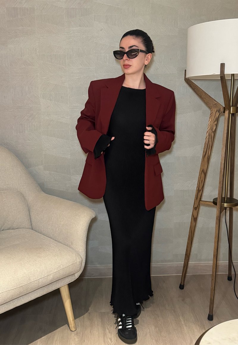 Blazer oversize bordeaux sur une robe midi noire à côtes, associé à des baskets noires. L'arrière-plan présente un mur de couleur claire et une lampe.