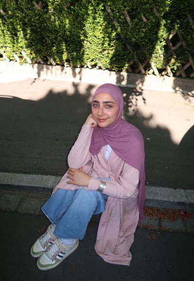 Una persona sentada en la acera vistiendo una camisa larga de color rosa claro, un hijab a juego y unos vaqueros anchos azules con deportivas verdes y beig.