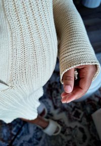 Light beige knitted sweater with ribbed texture and wide sleeves, worn with a partially visible hand and soft white slippers on a patterned rug.