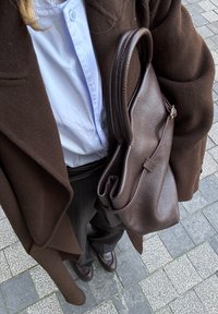 Personne portant un manteau marron, une chemise bleu clair boutonnée, un pantalon foncé et des chaussures, tenant un grand sac à main en cuir marron foncé sur un pavement carrelé.