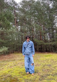 Giacca di jeans e jeans a gamba larga in blu chiaro, con un ginocchio strappato, abbinati a sandali marroni. Ambientato in un'area boschiva con muschio verde.