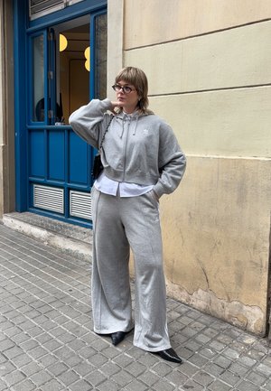 Sudadera gris y pantalones de chándal de pierna ancha combinados con una camisa blanca debajo. Zapatos negros de punta. El fondo presenta una puerta azul y una pared beige.