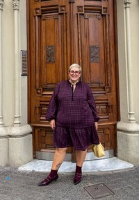 Karo haljina boje marune s volanima na rubu, dugim rukavima i mašnom oko vrata; uparena sa bordo čarapama i cipelama s oštrim noktima. Bež torba.