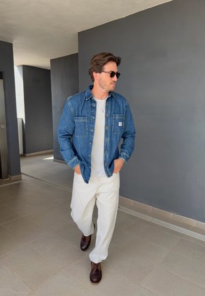 Hombre con gafas de sol, chaqueta vaquera azul, camisa blanca y pantalones blancos caminando en interiores con las manos en los bolsillos contra una pared gris de fondo.