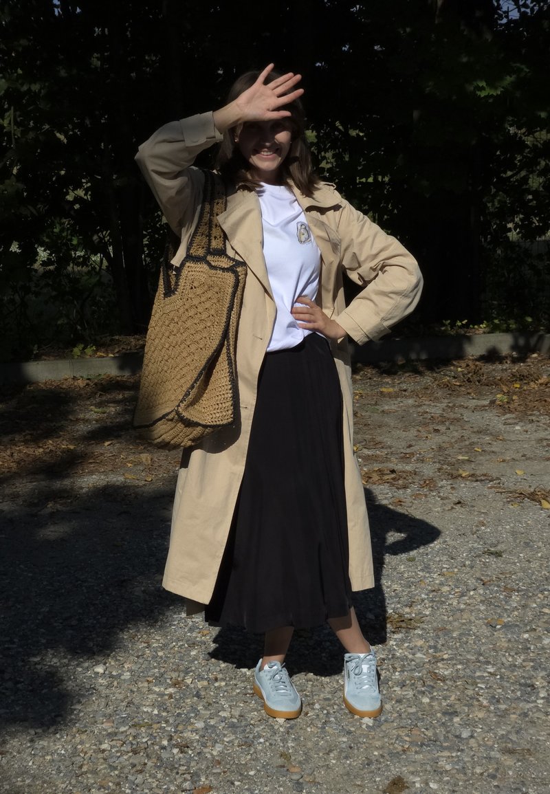 Beige παλτό τρέντς πάνω από άσπρη μπλούζα με λογότυπο, μαύρη φούστα και ανοιχτό μπλε αθλητικά με καφέ σόλες. Υφασμάτινη τσάντα με μαύρες λεπτομέρειες.