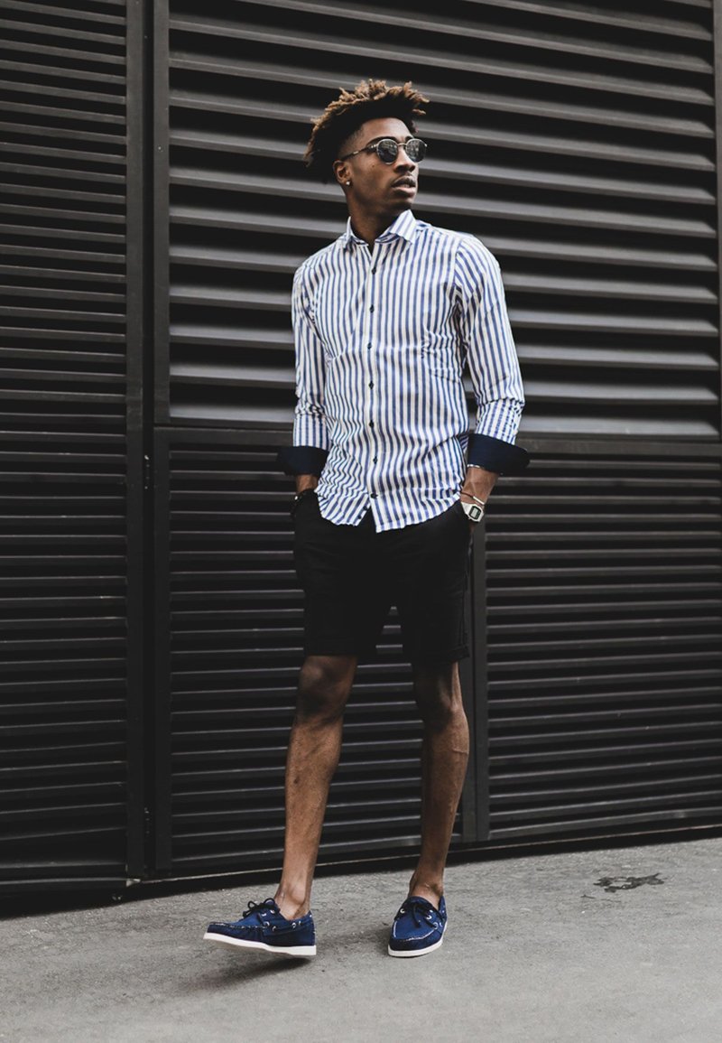 Rayé bleu et blanc, chemise à manches longues, shorts noirs et baskets bleues. Des lunettes de soleil et une montre sont portées. Se tient devant un fond à lattes horizontales.