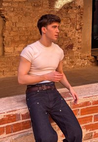 Hvid T-shirt med korte ærmer, mørke jeans med synlige sømme og et brunt bælte med logo. Sidder op ad en tekstureret murstensmur.