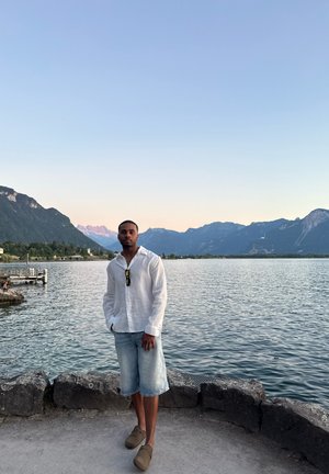 Camisa blanca de lino con botones, pantalones cortos de mezclilla claros, zapatos de slip-on marrones; de pie junto a un lago con montañas al fondo.