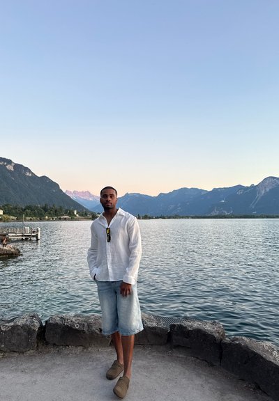 Camisa blanca de lino con botones, pantalones cortos de mezclilla claros, zapatos de slip-on marrones; de pie junto a un lago con montañas al fondo.