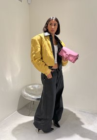 Femme en veste courte jaune, crop top blanc, pantalon large noir et talons noirs tenant un sac à main rose dans une pièce minimaliste avec une chaise en métal.