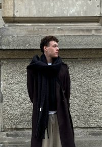 Sciarpa nera e lungo cappotto scuro indossato sopra una maglietta nera e pantaloni beige. Lo sfondo presenta una texture di parete in pietra grezza.