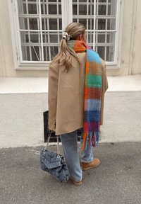 Beige oversized frakk over blå denim, fargerik stripete ullskjerf med frynser, grå quilted håndveske, og brune støvler på en bakgrunn av fortau.