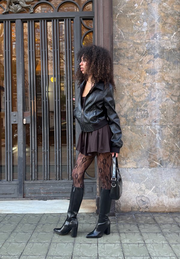 Sieviete ar cirtainiem matiem, valkājot melnu ādas jaku, īsu svārku, mežģīņu zeķubikses un ceļgalu augstus zābakus, stāv blakus dzelzs vārtu sētai, turot melnu somiņu.