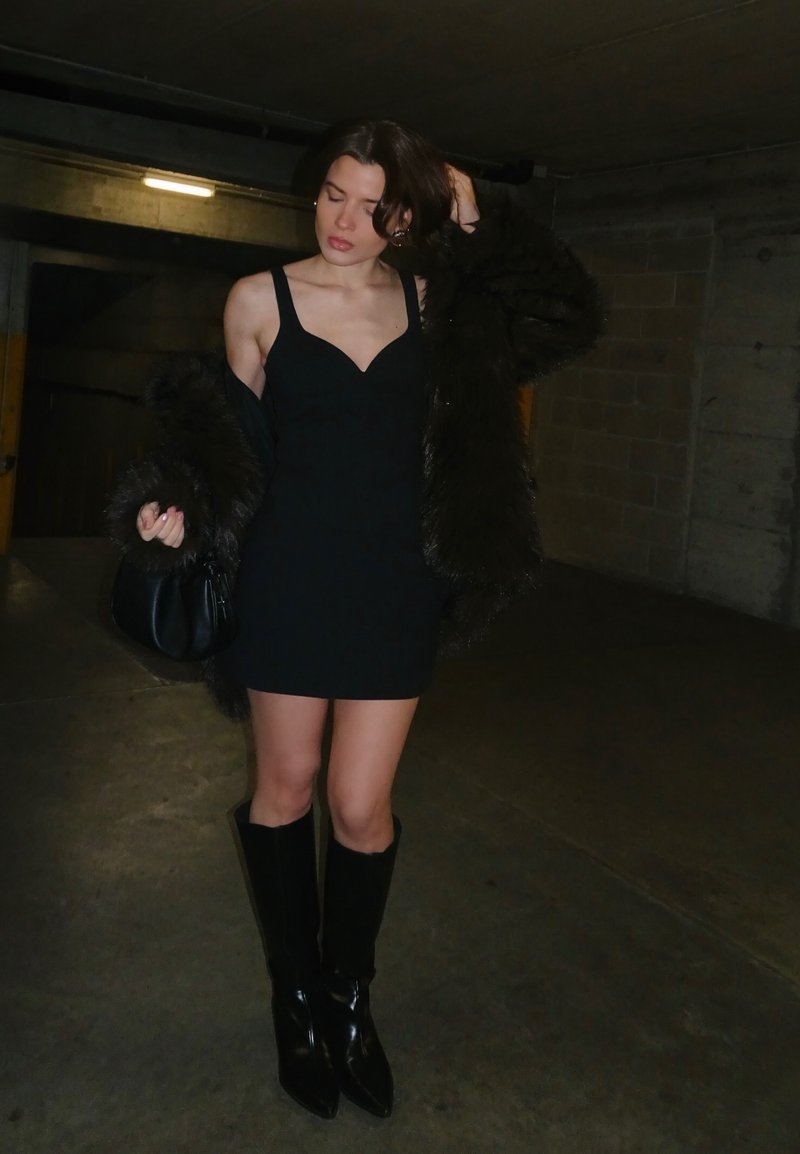 Femme en mini-robe noire et bottes, ajustant ses cheveux tout en portant un manteau en fourrure noire dans un parking souterrain faiblement éclairé.