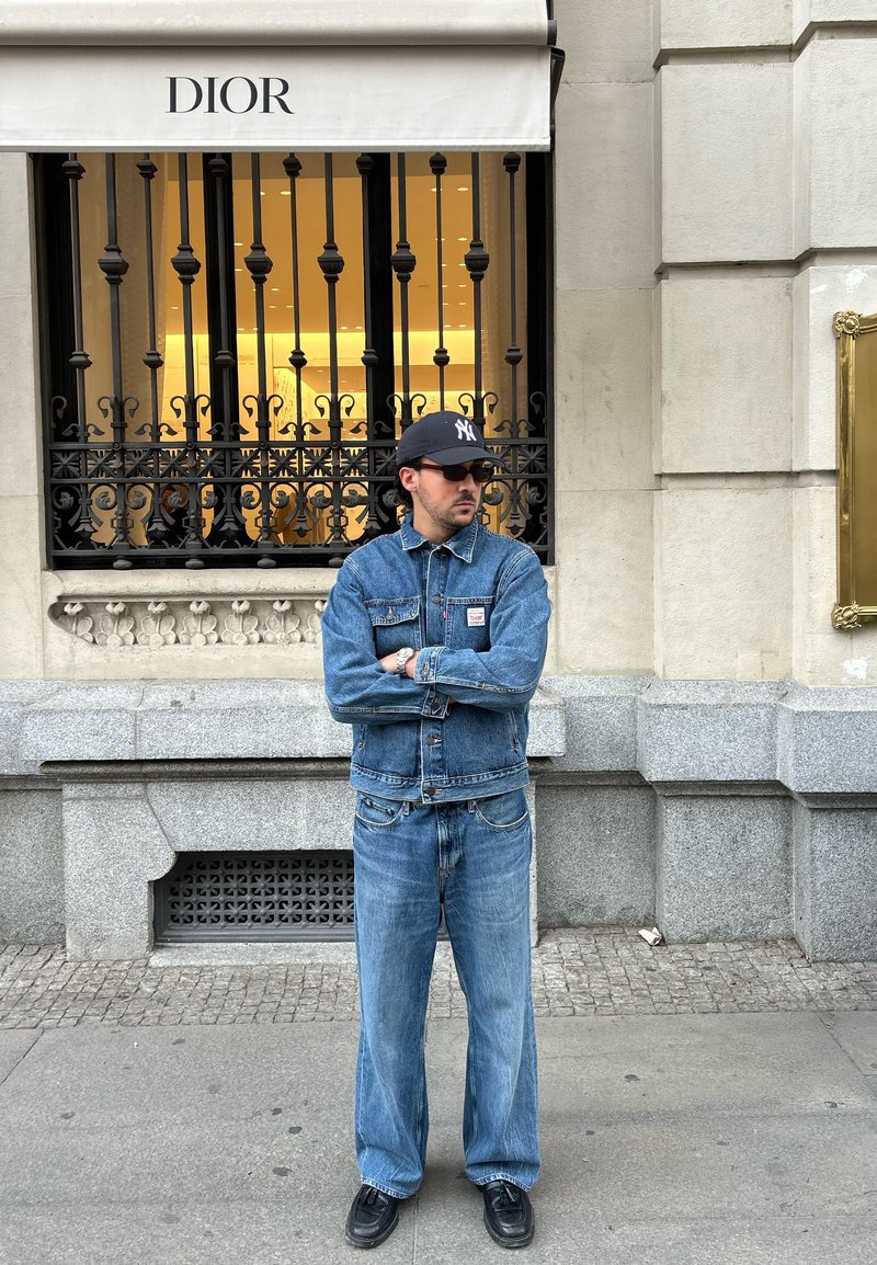 Veste en denim et jeans évasés en bleu clair, associés à des chaussures noires. L'endroit présente une vitrine Dior avec des barreaux ornés.