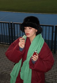 Κόκκινο φουσκωτό παλτό, πράσινος υφασμάτινος σκούφος, μαύρο καπέλο από γούνα. Κρατάει λιπ γκλος και κραγιόν. Μαλακό φωτισμό, εξωτερικός χώρος με μπλε διάδρομο.