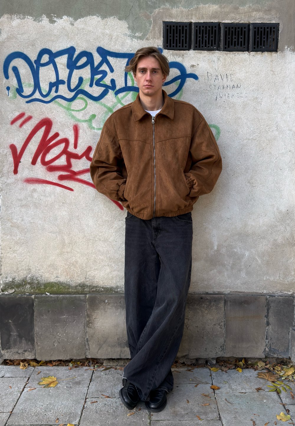 Hnědá semišová bunda bomber se zipem vpředu, v kombinaci s volnými černými džínami a černými botami, stojící před graffiti zdí.