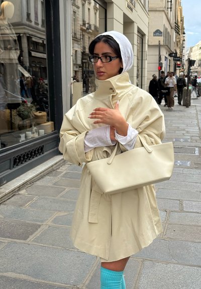 Mujer con abrigo beige y pañuelo blanco en la cabeza, de pie en la acera de la ciudad sosteniendo un bolso beige, con calcetines azules visibles y personas caminando de fondo.