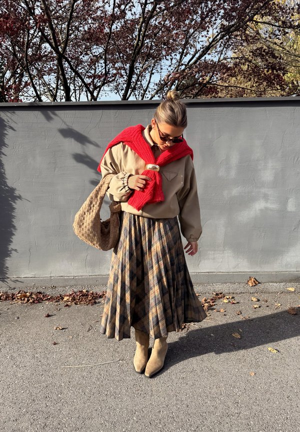 Beige oversized jumper with embroidered detail, red knitted scarf, checked pleated skirt in browns and yellows, tan ankle boots, woven bag.