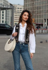 Črn vezen jopič preko bele srajce, v kombinaciji z visokim modrim kavbojkam. Velika krema torba z izrezanim ročajem. Mesto v ozadju.