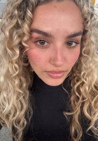 Nærbilde av en person med krøllete, blondt hår og naturlig sminke. De har på seg en svart høyhalsgenser og har glansfulle lepper. Brune øyne er synlige.