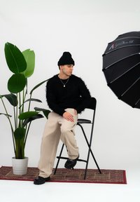 Pulover negru pufos, pantaloni bej cu croială largă, căciulă neagră, pantofi negri; așezată lângă frunze mari verzi și un reflector umbrelă negru.