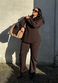 Brun blazer og matchende brede bukser lavet af glat stof, parret med en struktureret brun håndtaske og overdimensionerede røde solbriller.