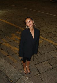 Svart, løst sittende jakke med lange ermer og belte i midjen, kombinert med svarte leggings og flip-flops, står på en steinoverflate.