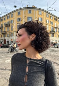 Maglione grigio con texture e dettagli a bottone sul colletto; modello con capelli ricci scuri, sfondo urbano con architettura gialla.