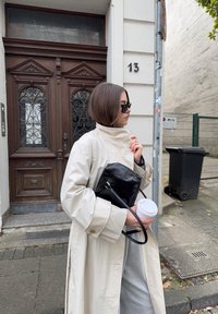 Beige trenčkot laiadest varrukatest, must käsip ключ sekä valge kohvitass käes. Taustal on ornamenteeritud puidust uks ja prügikast.