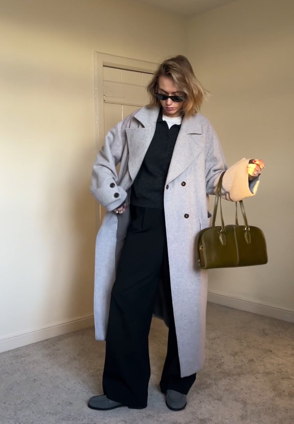 Person wearing an oversized light grey coat, black wide-leg trousers, dark sunglasses, and holding an olive green handbag in a neutral indoor setting.