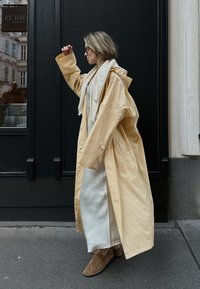 Beige Regenmantel mit oversized Schnitt und Kapuze, über einem weißen Kleid getragen, kombiniert mit braunen Schuhen; glatte Textur und schlichtes Design.