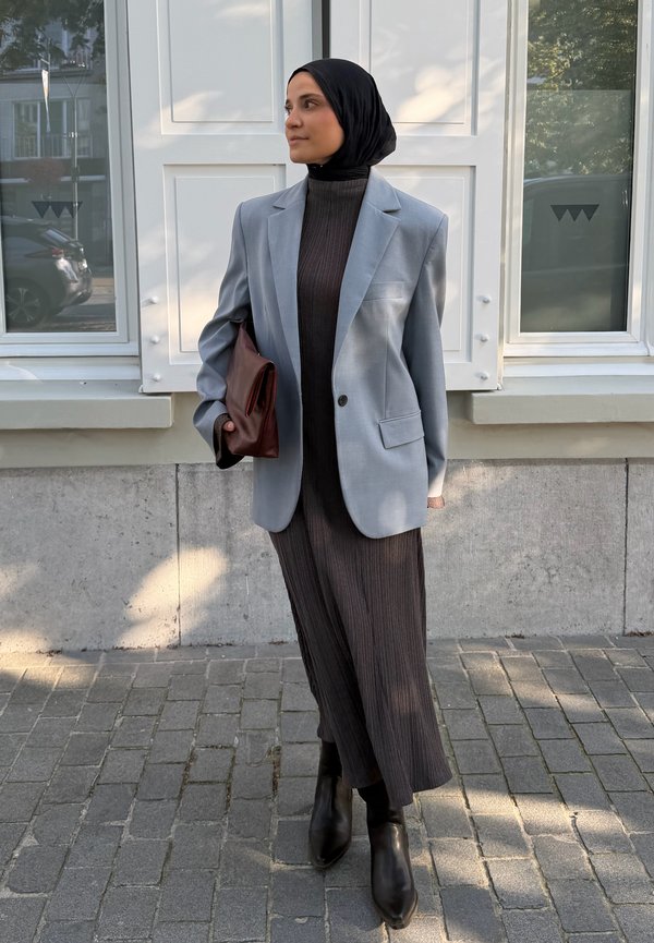Femme portant un hijab noir, un blazer gris, une robe plissée foncée, des bottes noires, tenant une pochette marron, debout dans une rue pavée devant un bâtiment blanc.