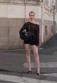 Przesadny brązowy sweter z dzianiny z przetarciami, w połączeniu z czarnymi szortami. Dodatki to czarna torebka i czarne szpilki z złotymi akcentami.