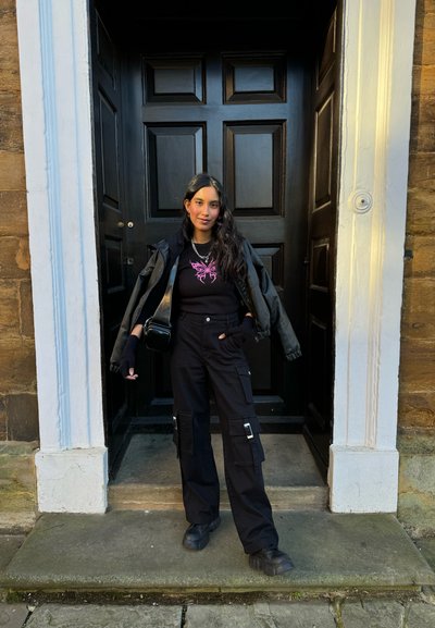 Pantalones cargo negros con bolsillos laterales, una camiseta negra con un diseño de mariposa rosa y una chaqueta negra de gran tamaño, de pie sobre escalones de piedra.