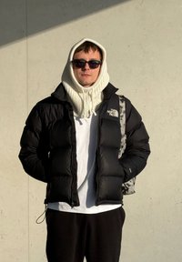 Jeune homme portant une doudoune noire de North Face, un capuchon en tricot blanc, des lunettes de soleil, une chemise blanche, et un sac à dos à motif camouflage contre un mur beige.