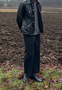 Personne portant une veste en cuir noire, un pantalon large noir et des chaussures noires, debout sur l'herbe près d'un champ labouré avec une maison au loin.