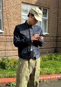 Blauwe spijkerjack met oranje stiksel, kaki cargobroek en een lichtgekleurde pet. Houdt een smartphone vast en draagt een apparaat rond de taille.